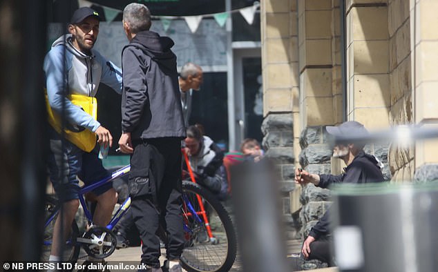 A man clutching a pipe sits on the ground as two men talk in front of him in Rawson Square