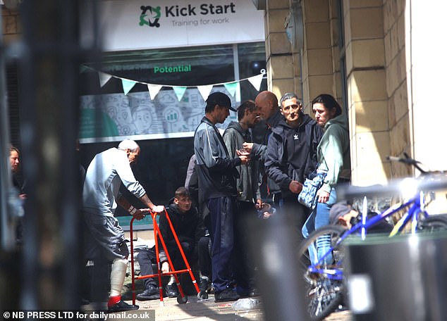 A man with a bandaged leg pushes a Zimmer frame towards a group of younger people holding a pipe and exchanging money