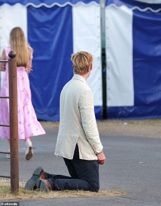 A suited male slumps to his knees on the grass at the Henley Regatta