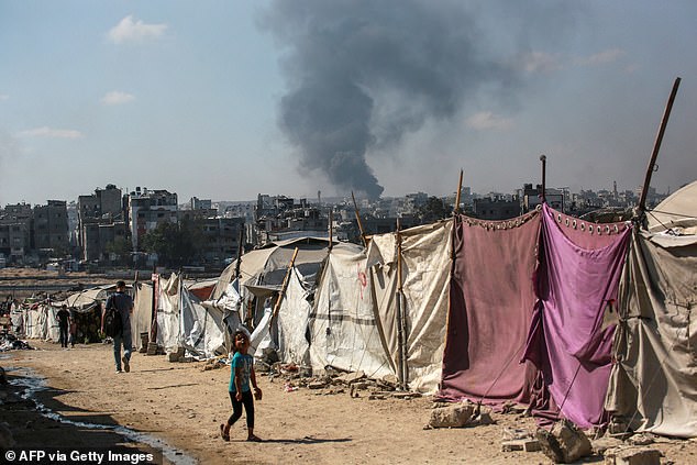 People walk past makesfift shelters as smoke billows east of Gaza City, in the central Gaza Strip during Israeli bombardment on July 6, 2025