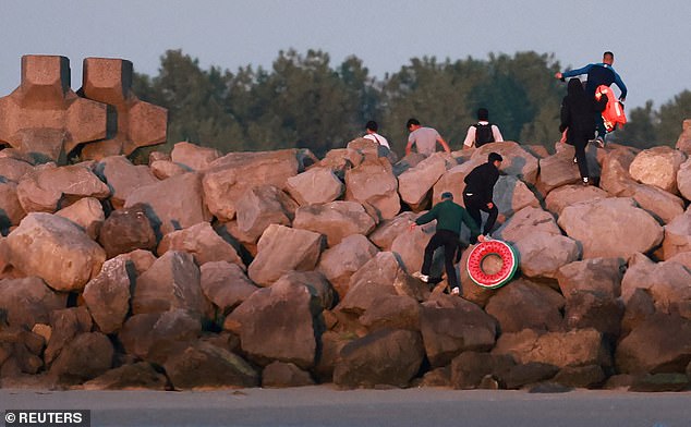 A group of migrants carrying life jackets and rubber rings are pictured as they climb over rocks after abandoning a dinghy whilst trying to cross the English Channel on a beach at Gravelines, France, this morning