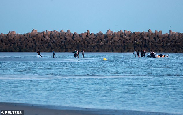A group of migrants carrying life jackets and rubber rings walk through the water after abandoning a dinghy whilst trying to cross the English Channel on a beach at Gravelines today