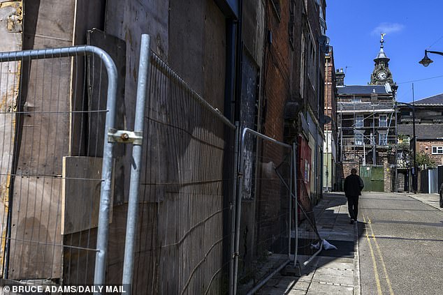 A boarded up building is surrounded by fencing, while a building at the end of the street can also be seen covered in scaffolding