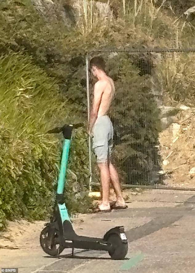 A male stands in plain sight and urinates near the beach huts with an e-scooter seen in the forefront
