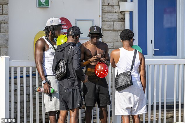 A group of males hang out on the seafront with balloons held to their lips. One of the males appears to be holding a gas canister