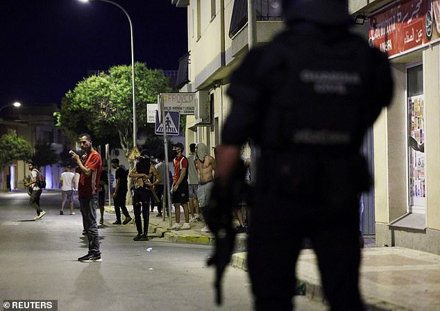 A man smokes as people gather on a street while a police officer keeps watch