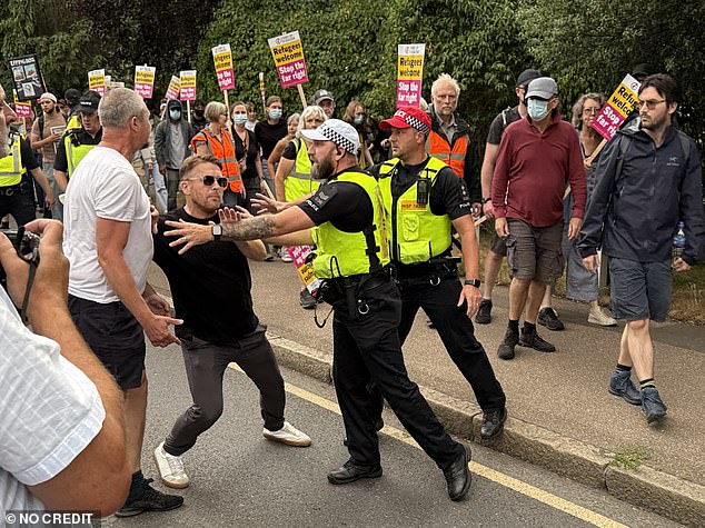A protestor is blocked by police as a counter-protest continues in the background, with people holding signs saying 'Refugees welcome, stop the far-right'