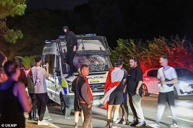 A man dressed in all black stands on the bonnet of a police van with its windows smashed