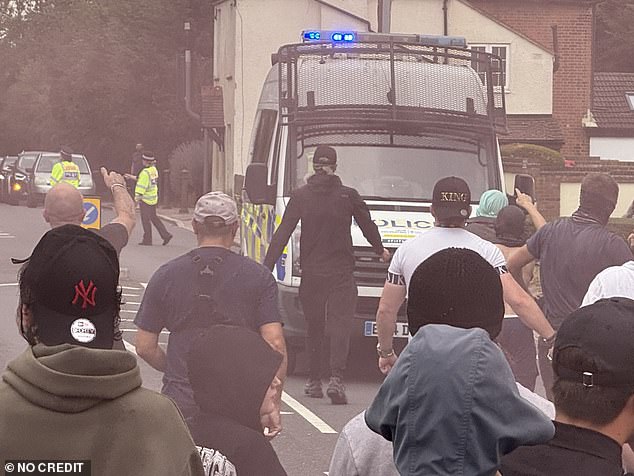 Another man dressed all in black approaches a police van as a braying mob watch on