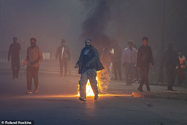 A group of men, some wearing masks, are seen at a distance from police with a fire blazing in the background