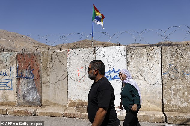 People on the Israeli side of the buffer zone walk past a Druze flag attached to one of the concrete barrier blocks, as they wait for new about relatives on the Syrian side