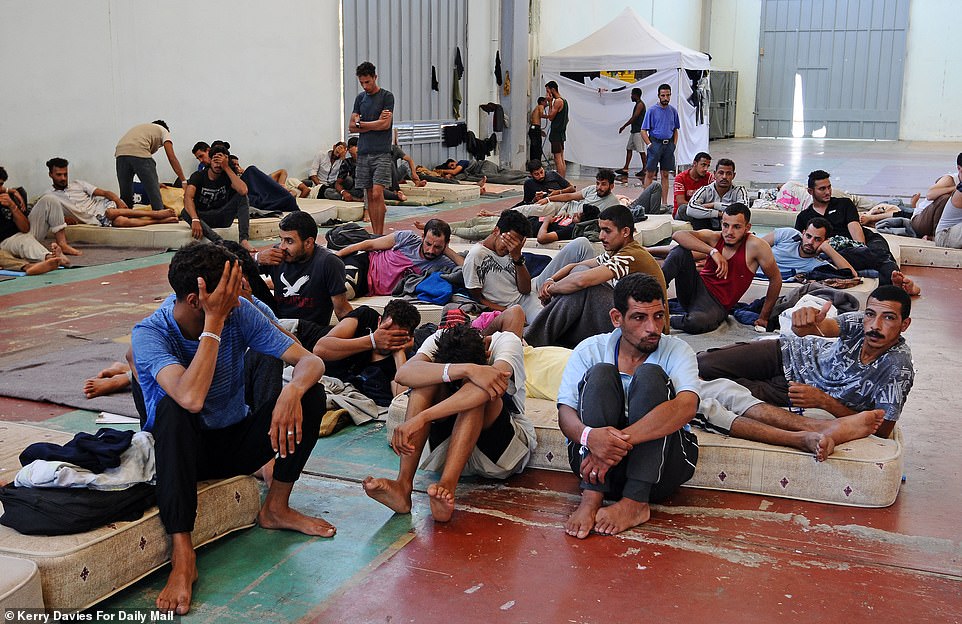 Migrants from Libya are guarded by armed police as they wait in stifling heat and sit on concrete floors at a warehouse in Chania town, Crete