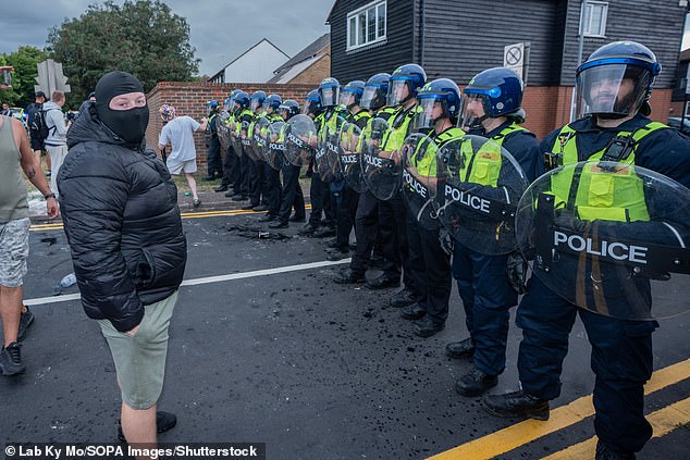 A masked protester faces a cordon of riot police officers preventing them from getting near a small group of pro-migrant protesters