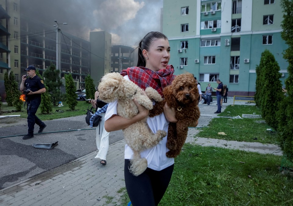 Woman carrying two dogs away from a damaged building.