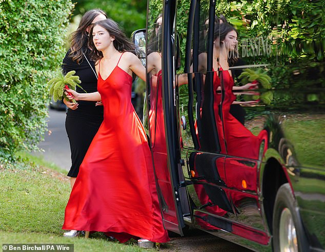 Another bridesmaid wearing a stunning red satin gown arrives at the church holding posies made of ferns