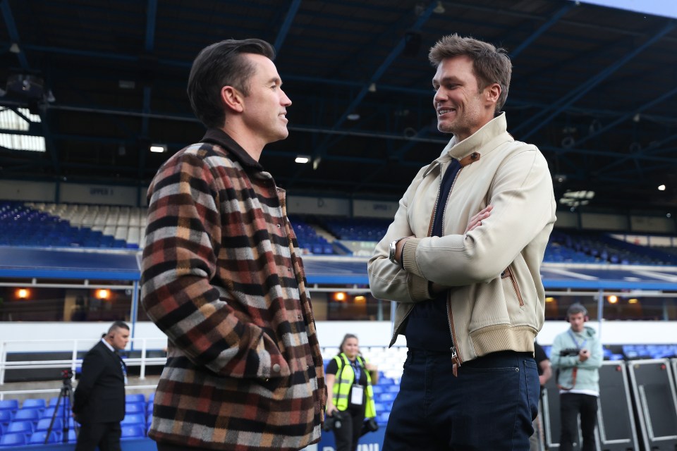 Rob McElhenney and Tom Brady talking at a football stadium.