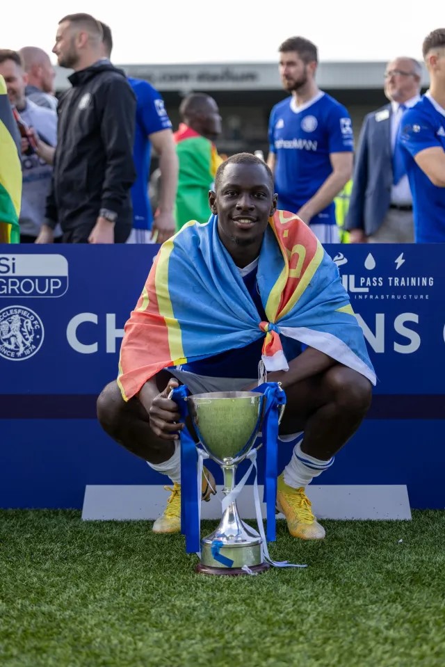 A soccer player crouching and holding a trophy, draped in a rainbow-colored flag.