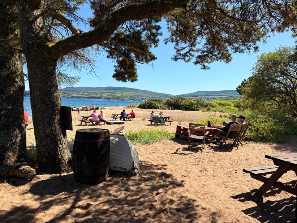 People sitting at tables on a beach with a view of the water and hills.
