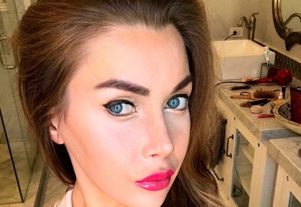 Close-up selfie of a woman with blue eyes and pink lipstick, a bathroom vanity in the background.