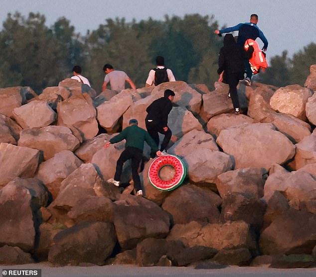 A group of migrants carrying life jackets and rubber rings climb over rocks after abandoning a dinghy whilst trying to cross the English Channel on a beach at Gravelines on Saturday