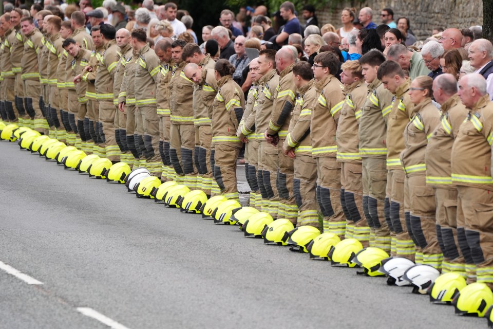 Firefighters and mourners lining a street before a funeral cortege.