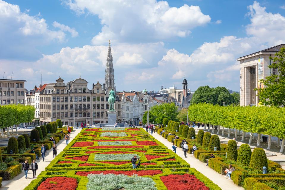 People strolling through a formal garden in Brussels, with buildings and the Town Hall tower in the background.