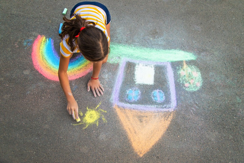 Child drawing a house and rainbow with chalk on asphalt.