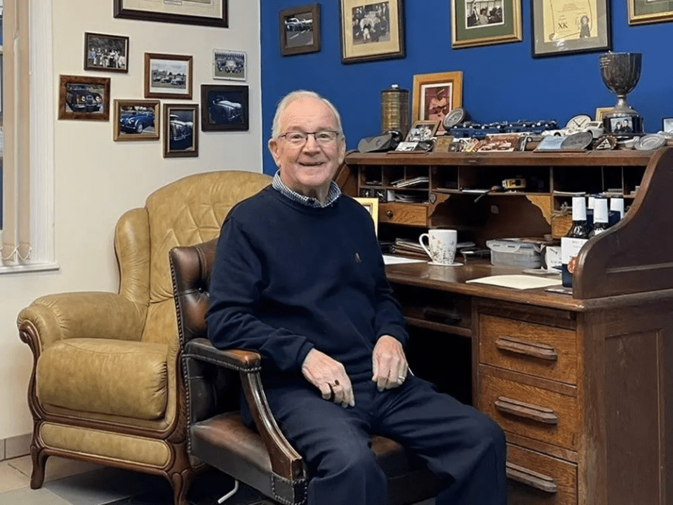 Man sitting in a leather armchair in front of a large wooden desk.