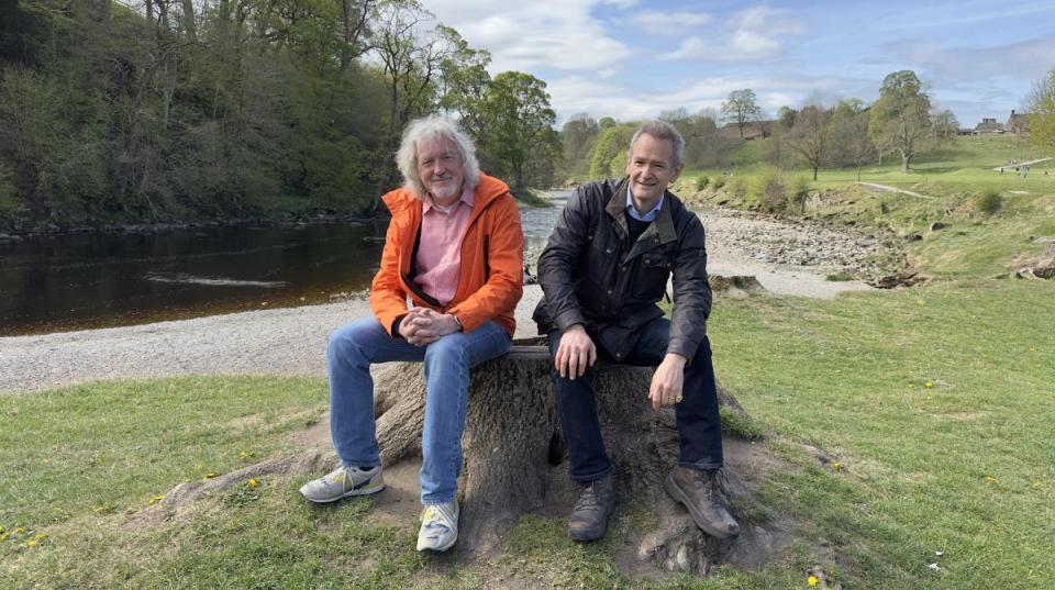 Alexander Armstrong and a companion sitting on a tree stump by a river.