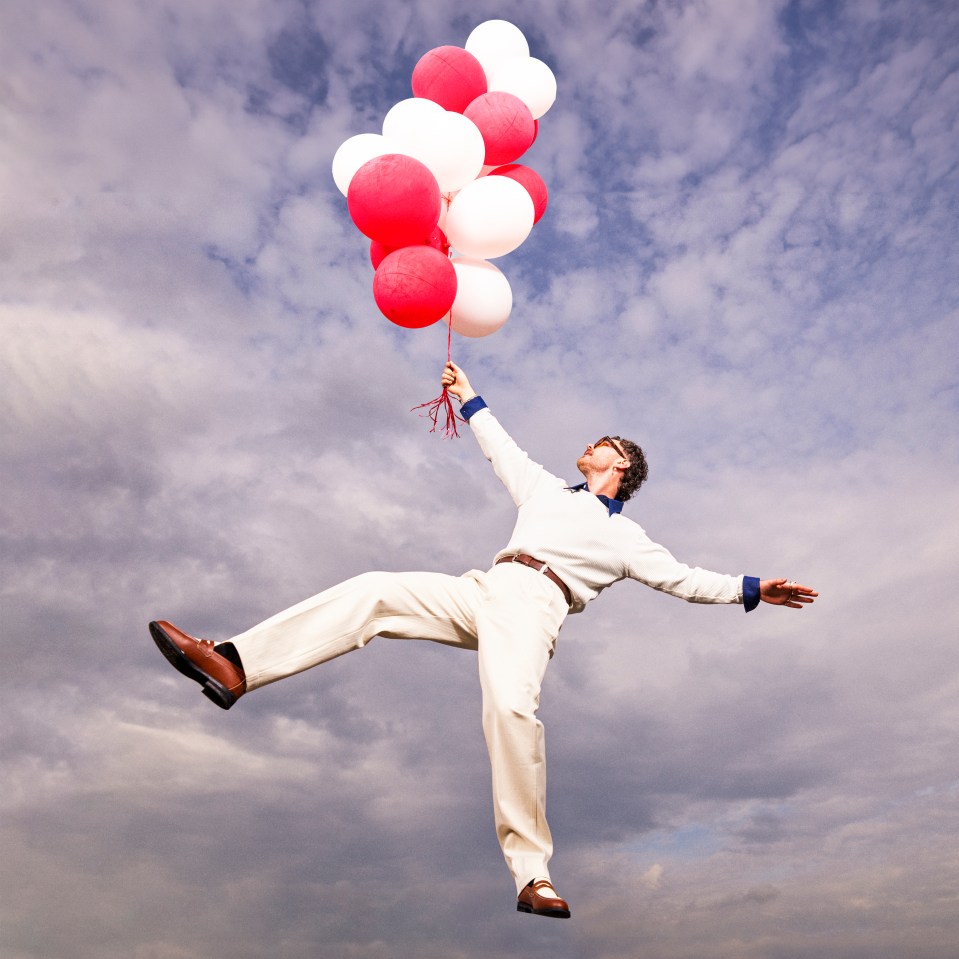 Man in white pants and shirt floats in the air holding red and white balloons.