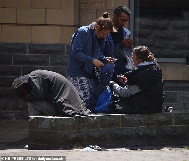 A group of people huddle round a woman with a pipe (right), as one man crouches over a wall covered with his coat, by businesses in the Rawson Square area of Bradford