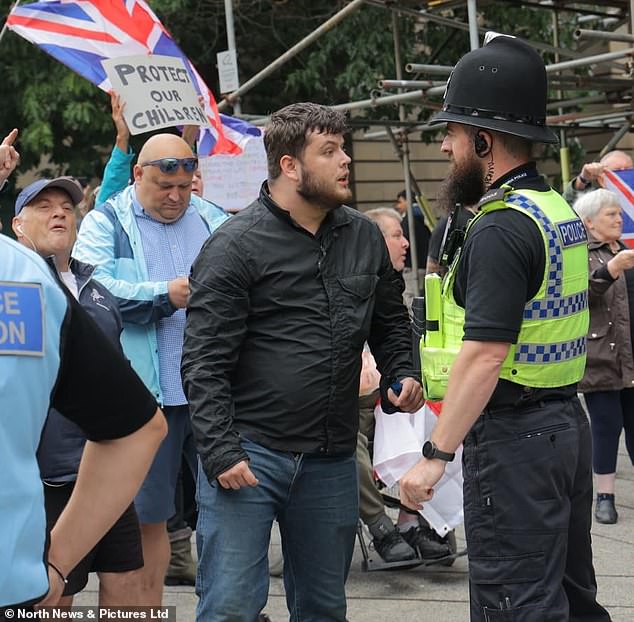 An anti-migrant activist in a black jacket clutches a vape while looking at a police officer