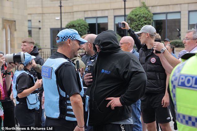 A man appearing to hide his face in a hood holds up his phone to a police liaison officer