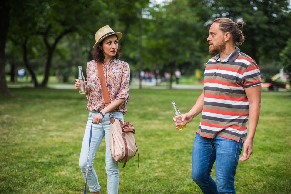 Couple walking a dog in a park, each holding a beer bottle.
