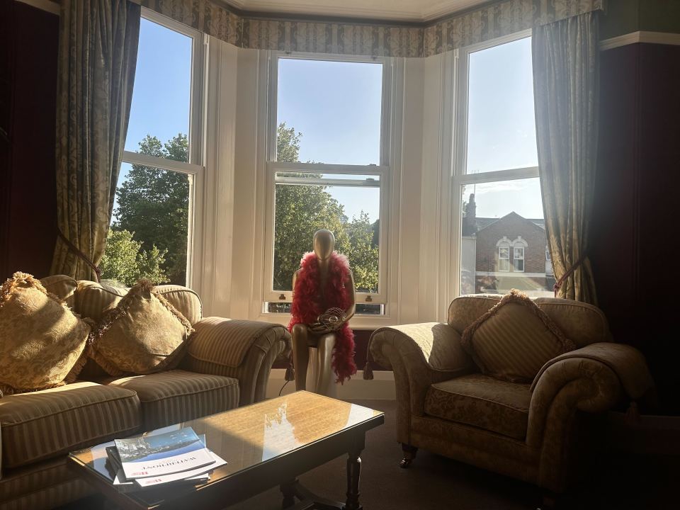 A mannequin wearing a red boa sits in a sunlit bay window of a B&B.