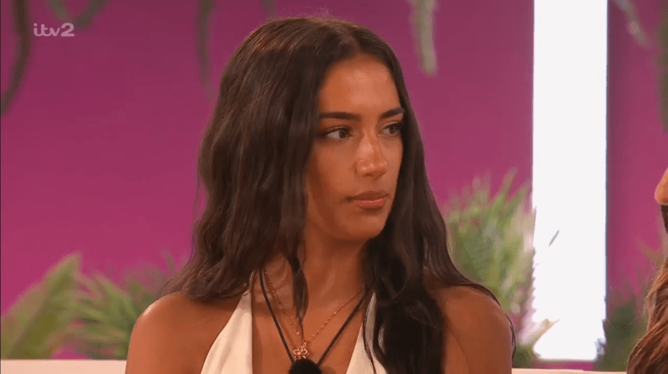 Screenshot of a woman with long brown hair, wearing a white halter top and necklace, looking pensive.