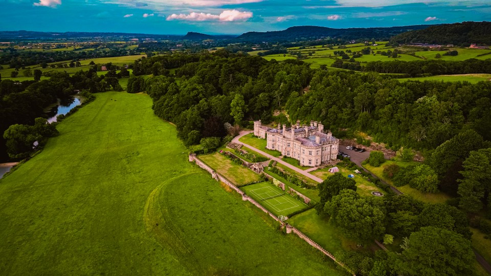 Aerial view of a large manor house with a tennis court and surrounding gardens.
