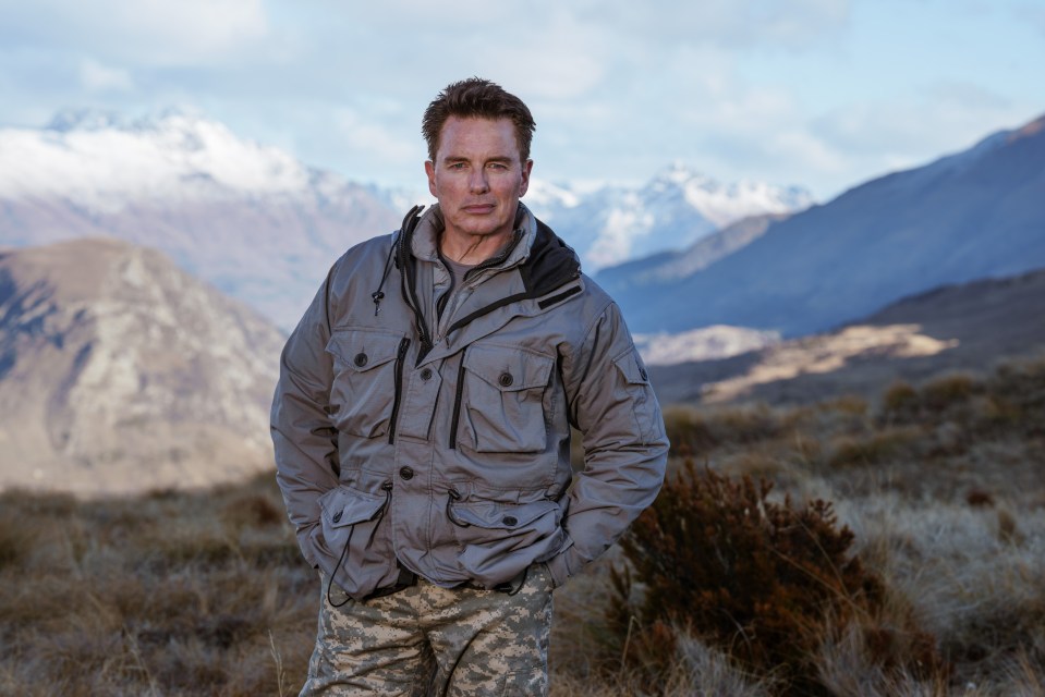 Portrait of John Barrowman in a gray jacket and camouflage pants, standing outdoors in front of a mountain range.