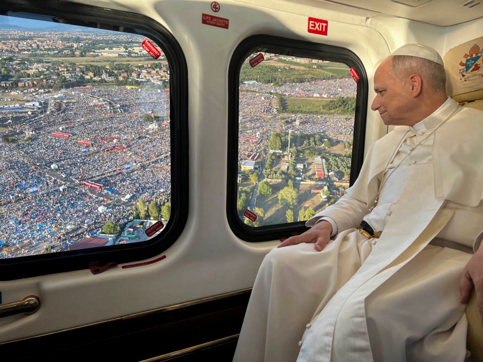 Pope Francis in a helicopter, looking out at a large crowd of people.
