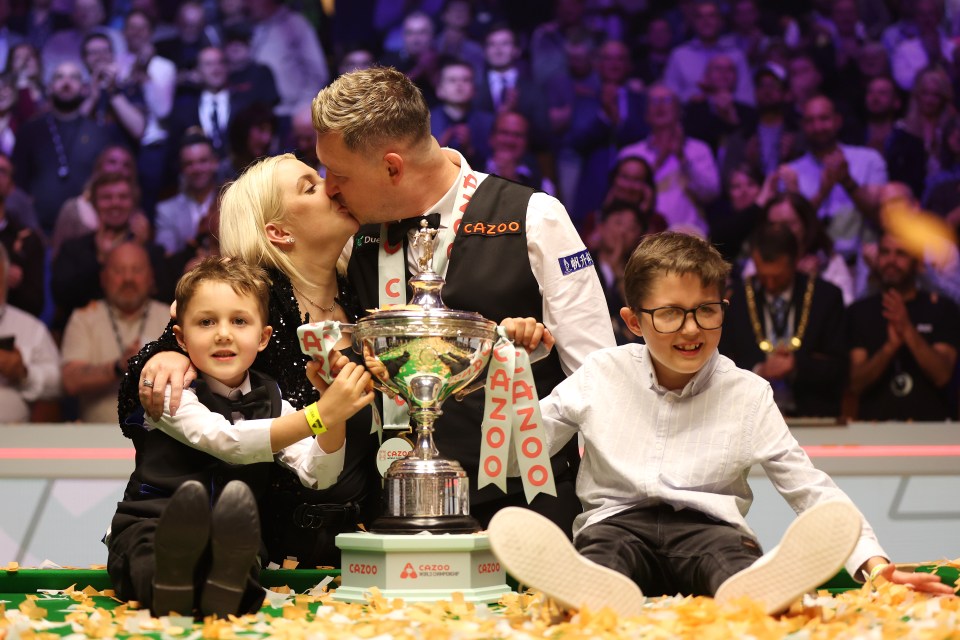 Snooker player Kyren Wilson kisses his wife while holding a trophy with his family.