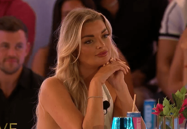 Screenshot of a blonde woman sitting at a table, looking pensive.