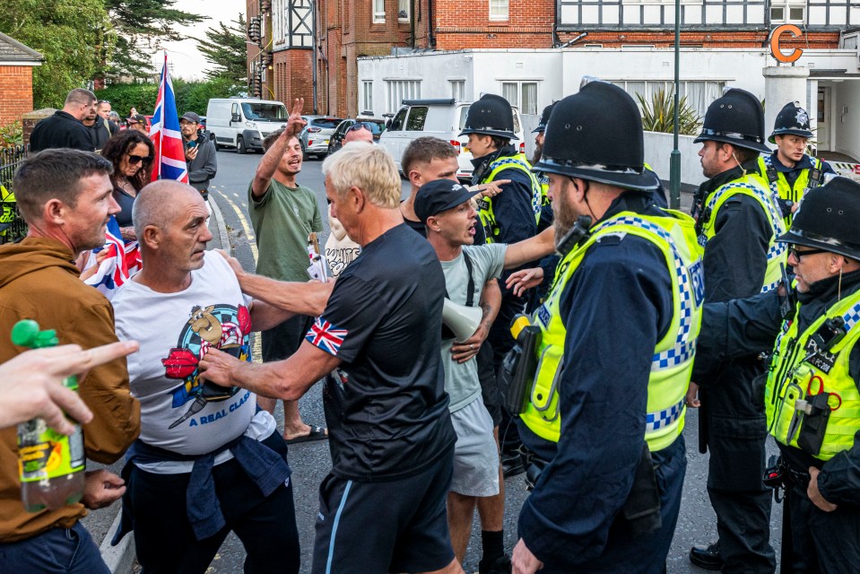 Protest outside a hotel; an asylum seeker flips off protesters.