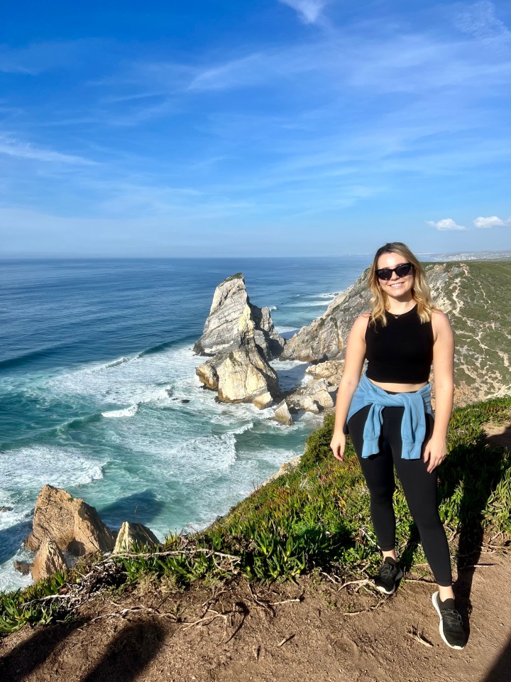 A woman is standing on the edge of a cliff, looking out at a beautiful ocean view.