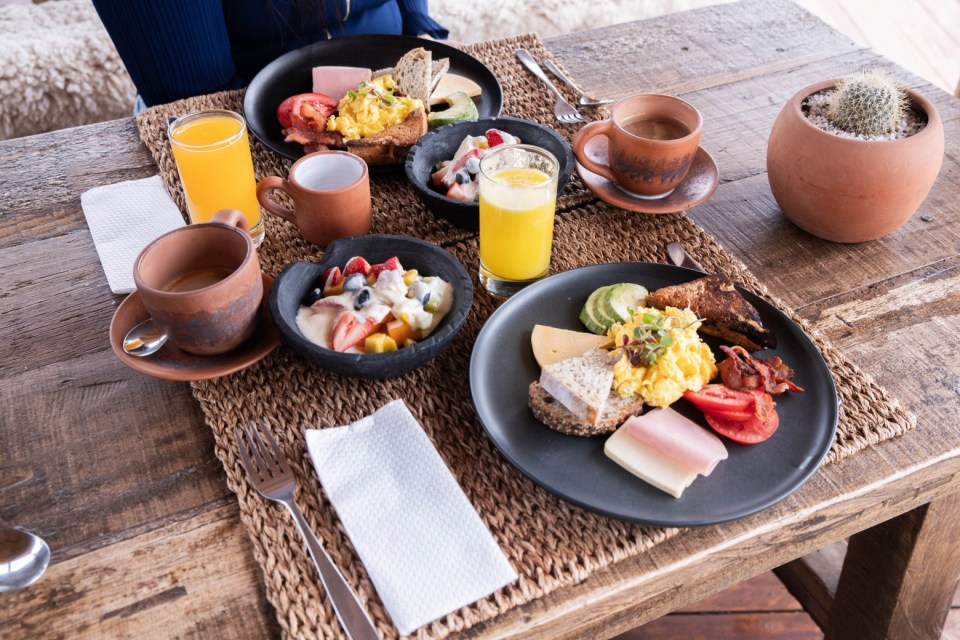 A beautifully arranged breakfast spread featuring scrambled eggs, fresh fruit, cheese, avocado, and toast is served on a rustic wooden table.