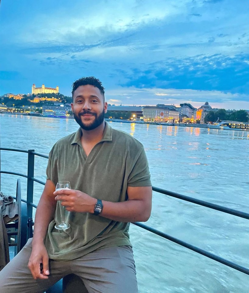 Man holding a drink on a boat with Buda Castle in the background.