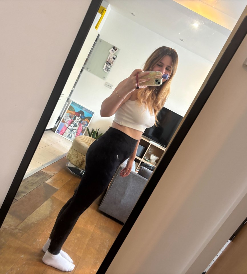 Woman in a white crop top and black leggings taking a mirror selfie.