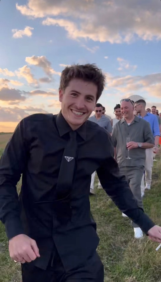 Man in black shirt smiling, walking towards the camera with a group of people in the background.