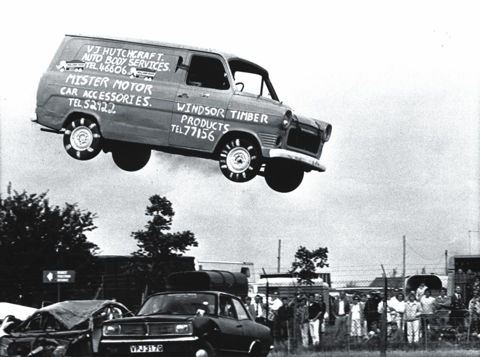 A van with advertisements painted on its sides airborne above a crowd of onlookers.