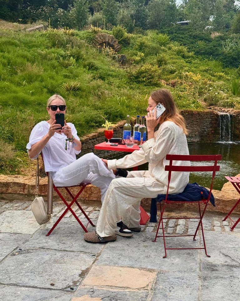 Two women sitting at a red table outdoors, one on a phone call, the other taking a photo.