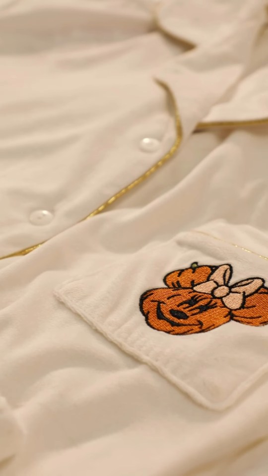 Close-up of a white pajama top with gold piping and a Minnie Mouse pumpkin patch embroidery on the pocket.